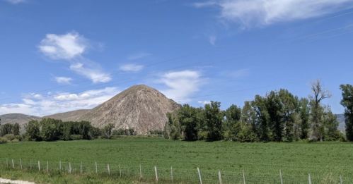 A grassy field with trees in the foreground and a large, rounded hill under a blue sky with scattered clouds.