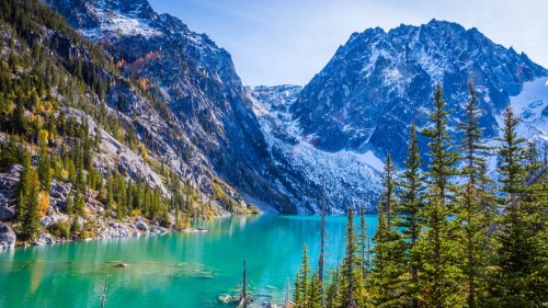 A serene mountain landscape featuring a turquoise lake, towering peaks, and lush evergreen trees under a clear blue sky.
