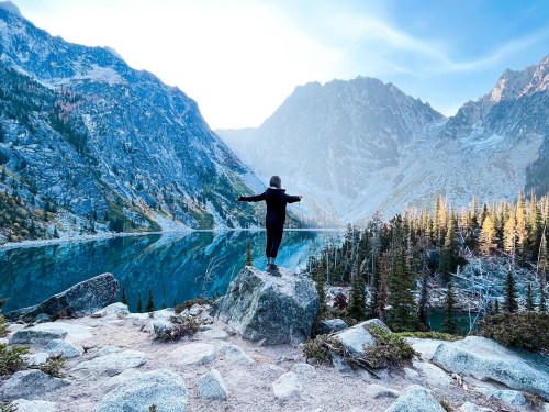 A person stands on a rock with arms outstretched, overlooking a serene lake surrounded by mountains and trees.