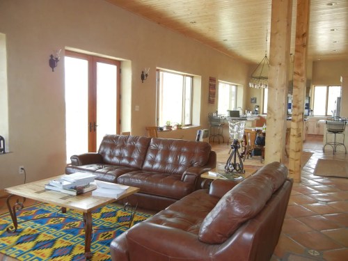 Cozy living room with leather sofas, wooden beams, and a colorful rug, leading to a kitchen area in the background.
