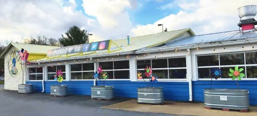 Colorful restaurant exterior with flower decorations, large windows, and a bright sign encouraging social media follows.
