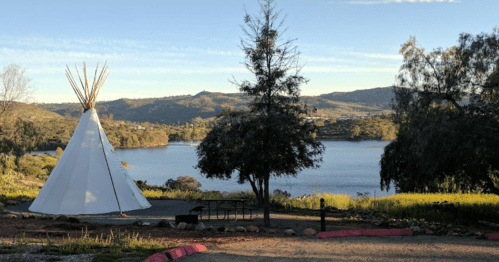 A white teepee stands near a lake, surrounded by trees and hills under a clear blue sky.