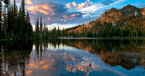 A serene lake reflects colorful clouds and trees, surrounded by mountains under a vibrant sunset sky.