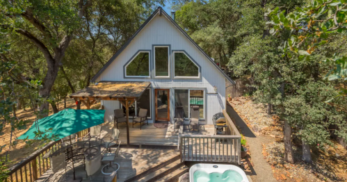 A modern A-frame cabin surrounded by trees, featuring a deck, outdoor seating, and a hot tub.