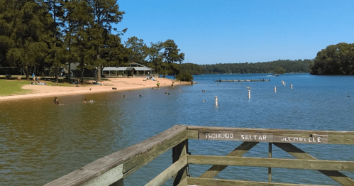 A serene lake scene with a sandy beach, people swimming, and a wooden pier under a clear blue sky.
