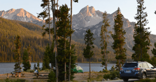 A serene lakeside scene with mountains in the background, trees, a tent, and a parked car.