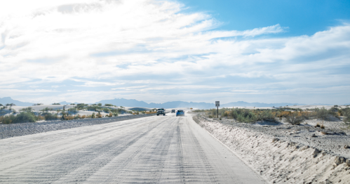A sandy road stretches into the distance, flanked by desert vegetation and mountains under a bright blue sky.