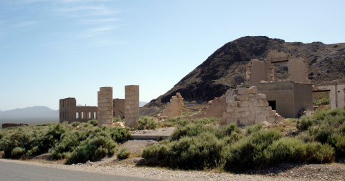 Ruins of a stone building surrounded by sparse vegetation and mountains under a clear blue sky.