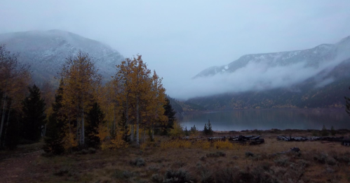 A serene lake surrounded by mountains, with autumn trees and misty clouds under a gray sky.