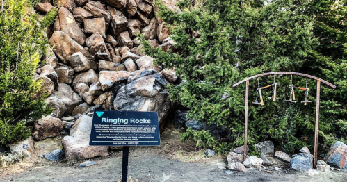 Sign for "Ringing Rocks" next to a rock formation and a metal frame with hanging bells in a natural setting.