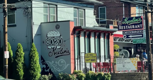 A restaurant named Kaelin's with a burger logo, outdoor seating, and a sign advertising cheeseburgers.