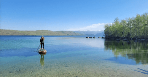 A person stands on a paddleboard in calm, clear water surrounded by trees and mountains under a blue sky.