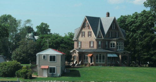 A large, historic brick house with a steep roof, accompanied by a smaller modern home on a green lawn.