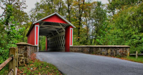 A red covered bridge with a stone wall entrance, surrounded by lush green trees and a paved road.