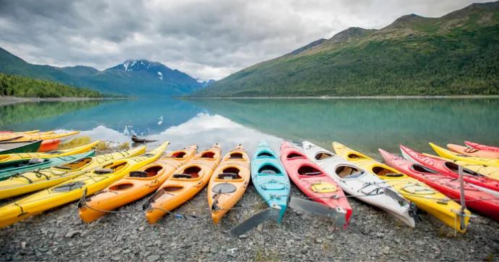 Colorful kayaks lined up on a rocky shore beside a calm lake, surrounded by mountains and cloudy skies.