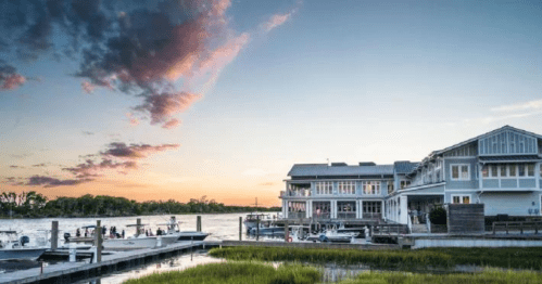 A serene waterfront scene at sunset, featuring a modern building and boats docked along a grassy shoreline.