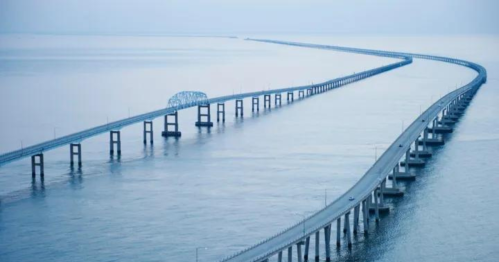 A long bridge stretches over calm water, disappearing into the horizon under a soft blue sky.