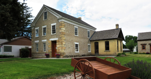 Historic stone house with a wooden wagon in the foreground, surrounded by green grass and trees under a cloudy sky.