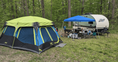 A green and black tent next to a camper under a blue canopy in a wooded campsite.