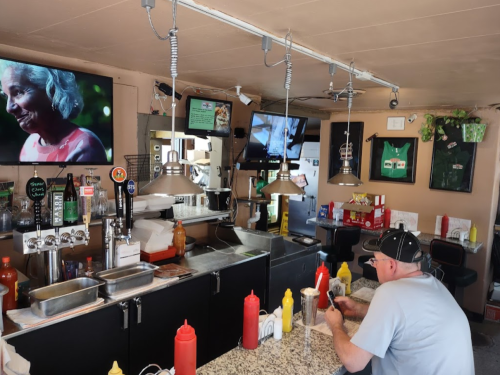 A diner with a man sitting at the counter, watching TV, surrounded by condiments and kitchen equipment.