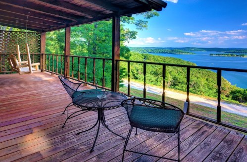 A scenic view from a wooden deck overlooking a lake, with two chairs and a small table on the porch.