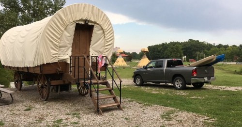 A covered wagon on wheels next to a pickup truck with a kayak, set in a grassy area with teepees in the background.