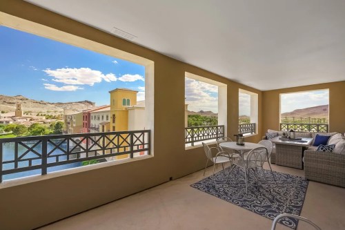 A cozy balcony with seating, overlooking a scenic view of buildings and mountains under a blue sky.