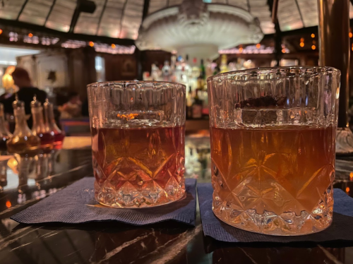 Two crystal glasses of amber whiskey on blue coasters, set on a dark marble table in a cozy bar atmosphere.