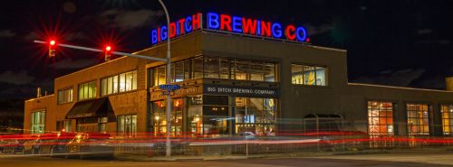 A brightly lit brewery at night with a neon sign reading "Big Ditch Brewing Co" and traffic lights in the foreground.