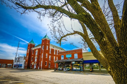 Historic red brick building with green spires, storefronts below, and a bare tree in the foreground against a blue sky.