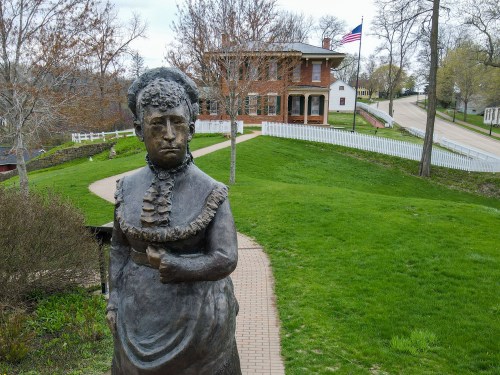 A bronze statue of a woman stands in a park, with a historic building and American flag in the background.