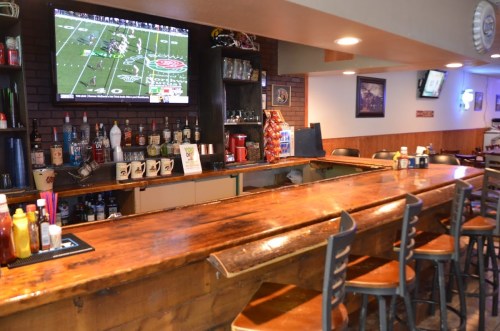 A cozy bar with a wooden counter, shelves of drinks, and a TV showing a football game in a casual setting.