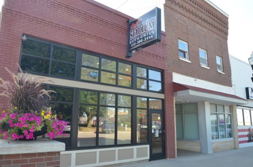 Exterior of a restaurant with a sign reading "Stadium Bar & Grill," featuring large windows and colorful flower planters.