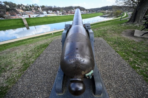 A historic cannon facing a river, with a scenic view of a town and greenery in the background.