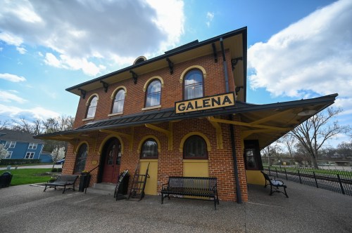 Historic Galena train station with brick exterior, yellow accents, and benches outside under a partly cloudy sky.
