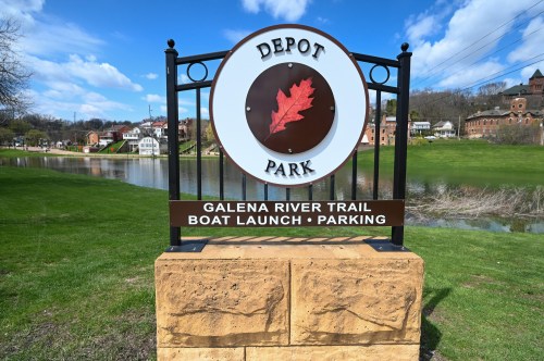 Sign for Depot Park featuring information about the Galena River Trail, boat launch, and parking. Scenic background.