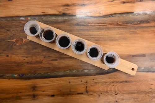 A wooden paddle holds six small cups filled with dark liquid, placed on a rustic wooden table.