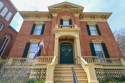 Historic brick building with green shutters, a decorative porch, and a flag in front, under a blue sky.