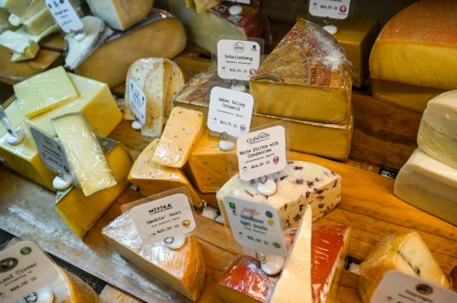 A variety of cheese blocks displayed on wooden shelves, each labeled with names and prices.