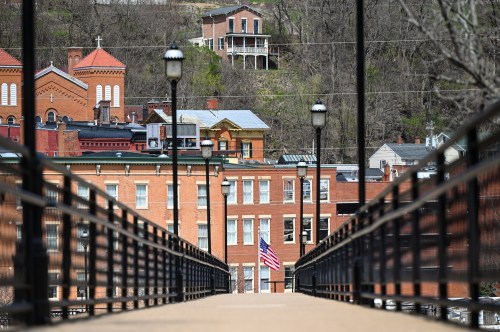 A view of a bridge leading to historic buildings, with an American flag and hillside homes in the background.