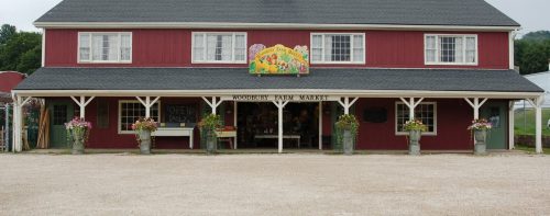 Red barn-style building with a sign reading "Woodbury Farm Market," surrounded by flower pots and gravel driveway.