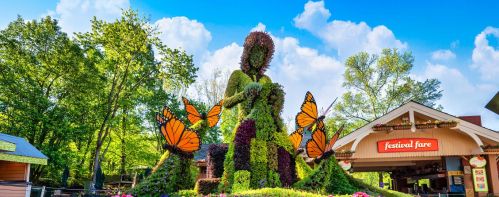 A large topiary figure surrounded by colorful butterflies, set against a bright blue sky and green trees.