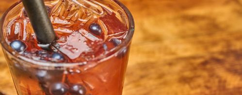 A close-up of a glass filled with a dark beverage, ice, and blueberries, with a straw resting inside.