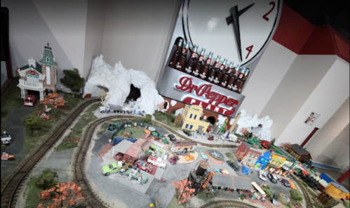 A miniature train set with buildings, trees, and a Dr Pepper sign, set against a clock backdrop.