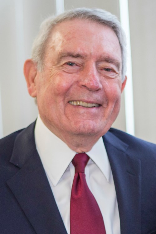 A smiling older man in a suit and tie, with gray hair, standing in front of light-colored vertical blinds.