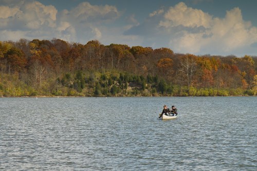 Two people in a small boat on a calm lake, surrounded by autumn-colored trees and a cloudy sky.