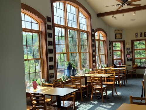 Bright, airy restaurant interior with large windows, wooden tables, and chairs, surrounded by greenery outside.