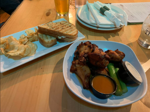 A plate of chicken wings with celery and dipping sauce, alongside a side of chips and toasted bread on a wooden table.