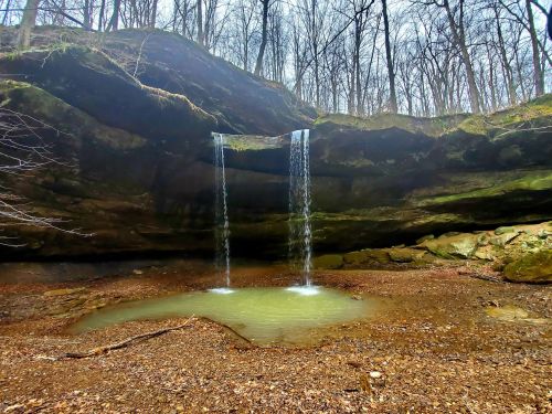 A serene waterfall cascades from a rocky overhang into a small pool, surrounded by bare trees and earthy terrain.
