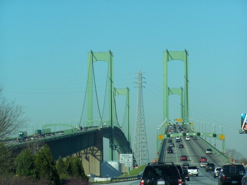 A green suspension bridge with traffic, set against a clear blue sky. Power lines are visible in the background.